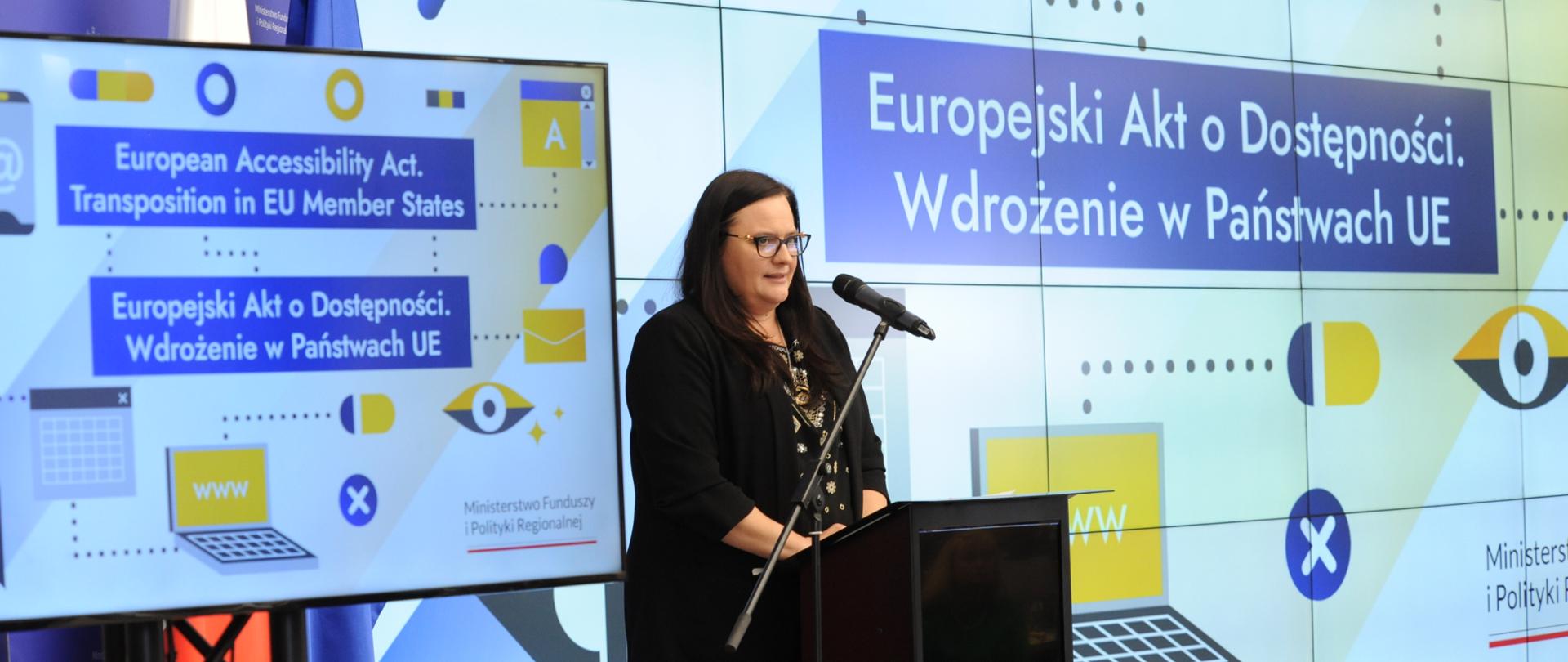 Zdjęcie przedstawia kobietę przemawiającą na konferencji związanej z wdrożeniem Europejskiego Aktu o Dostępności w państwach UE. W tle znajduje się duży ekran z napisami w języku polskim i angielskim dotyczącymi tego aktu oraz logotyp Ministerstwa Funduszy i Polityki Regionalnej. Kobieta stoi przy mównicy i mówi do mikrofonu, co sugeruje, że jest jednym z prelegentów tego wydarzenia.